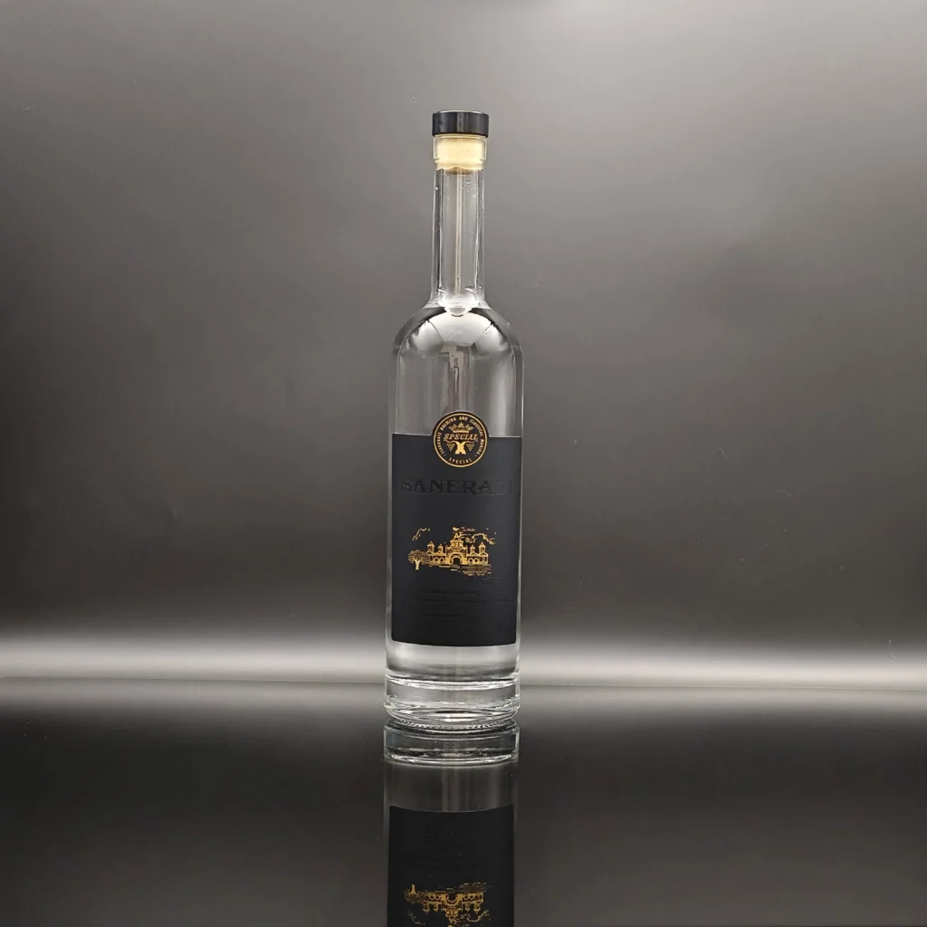 A classic cylinder-shaped glass vodka bottle made of crystal-clear super flint glass, featuring a heavy base, a natural T-cork closure, and a premium matte black paper label with elegant gold foil printed branding.