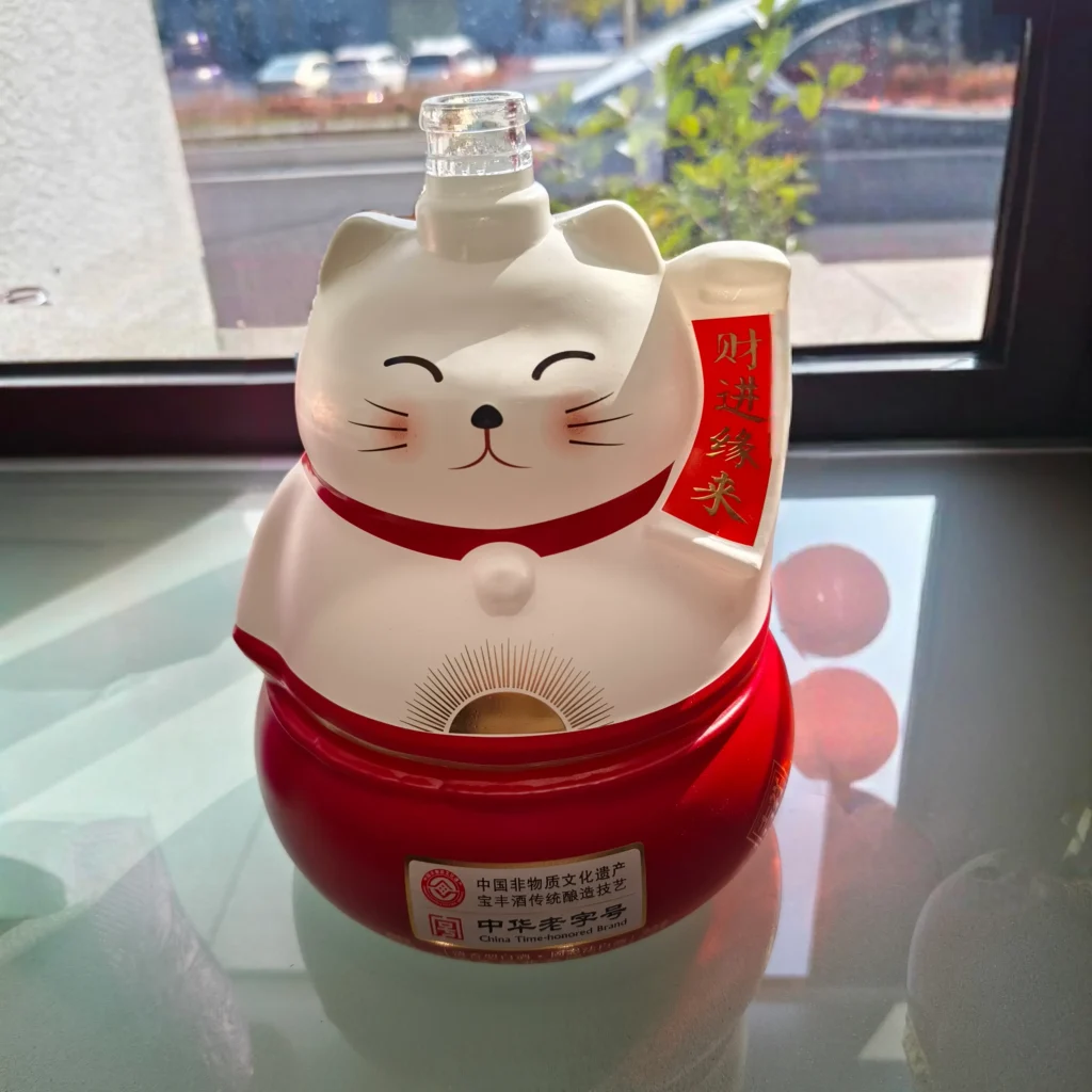 A custom lucky cat-shaped spirits bottle featuring a precise multi-color coating of matte white and vibrant red, decorated with fine facial expressions and metallic gold decal branding.