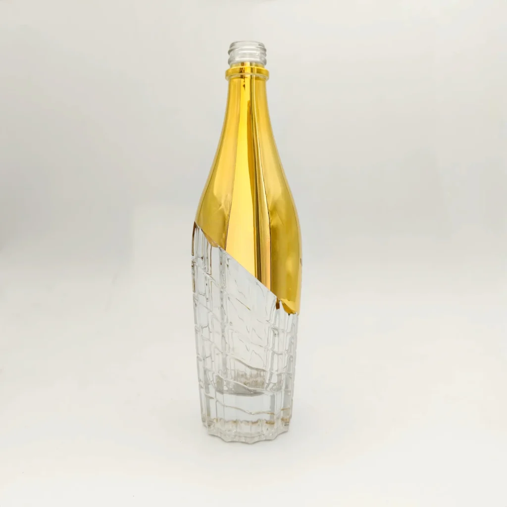 A premium spirits glass bottle showing a partial metallic gold electroplating finish on the upper body, separated by a sharp diagonal masking line from the clear glass base with an intricate geometric grid texture.