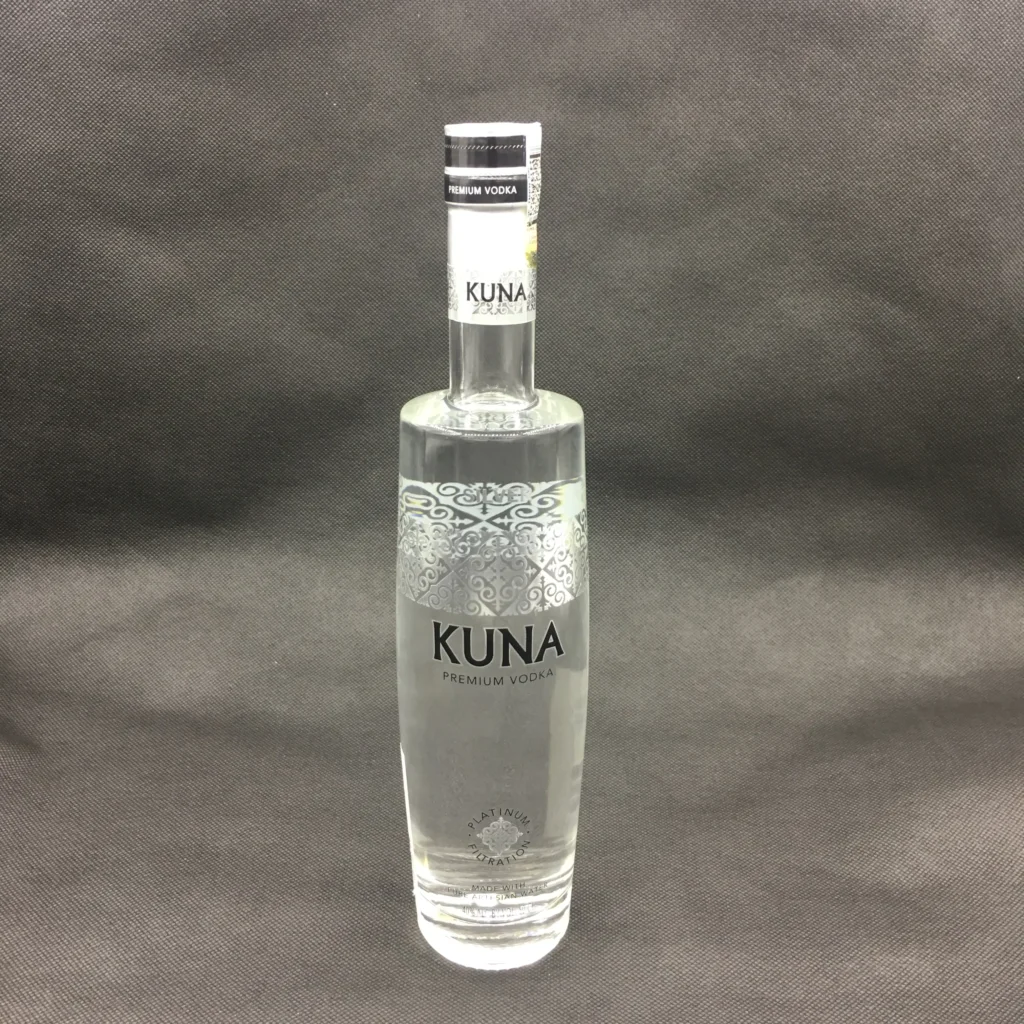 A tall, slim cylinder glass vodka bottle made of clear super flint glass, featuring an intricate silver filigree pattern and crisp black custom decal printing for Kuna Premium Vodka, finished with a heavy bottom.